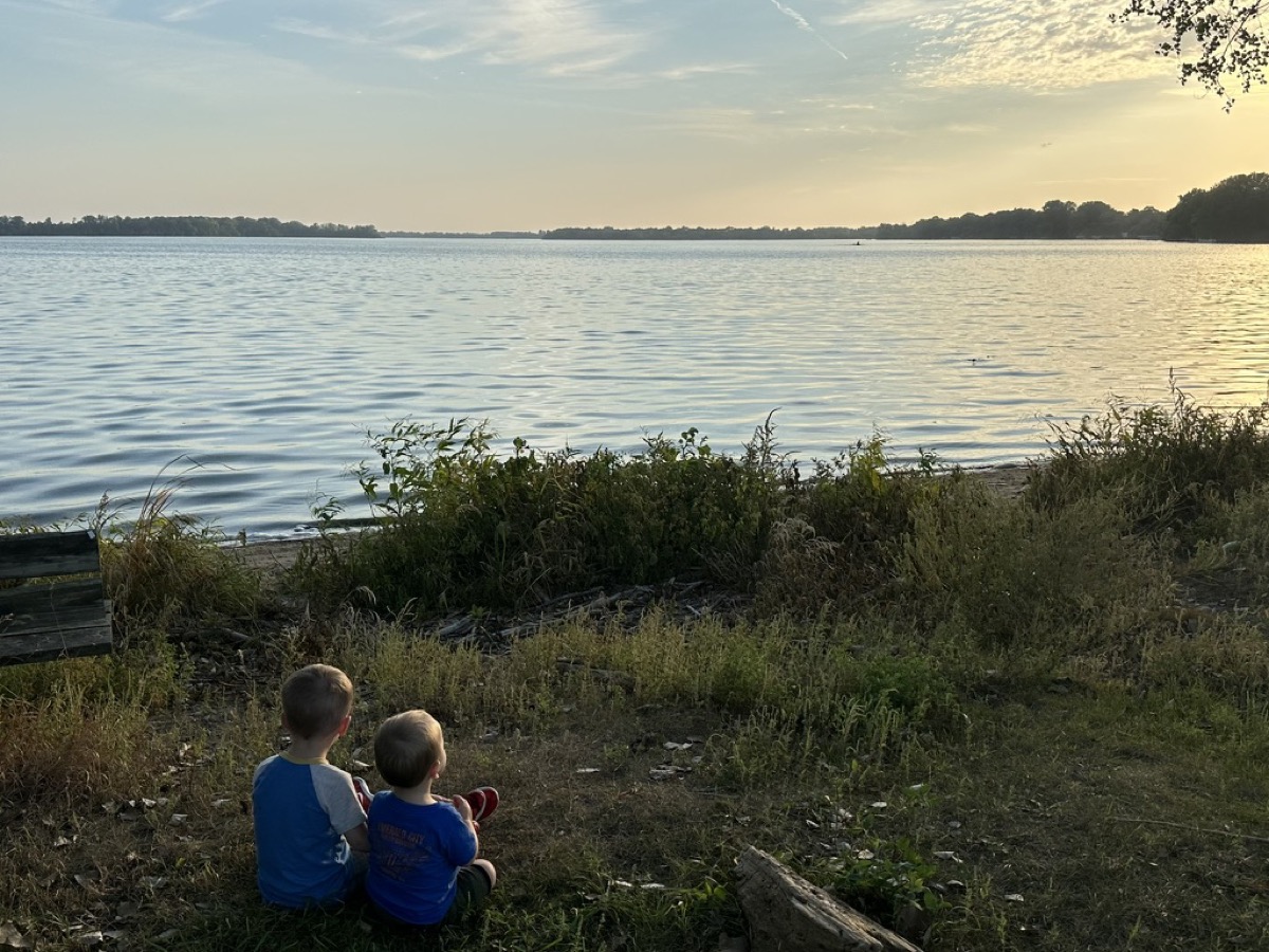 Two young boys sitting on the shore watching the lake at sunset